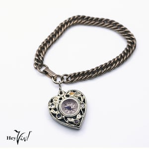 May include: Silver-toned bracelet with a heart-shaped pendant. The pendant features a compass design and is adorned with small, colorful gemstones. The bracelet has a chain link design and a clasp closure. The brand name "Hey Viv!" is visible in the bottom left corner.