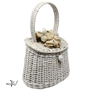 May include: White wicker basket with a curved handle and a decorative floral arrangement. The basket has a rounded shape and a small, round clasp. The basket is a light, neutral color.