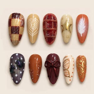 May include: A collection of ten artificial nails with various designs. The designs include a gold and brown plaid pattern, a white leaf design, a red grid pattern, a gold star, a white feather, and more. The nails are oval-shaped.