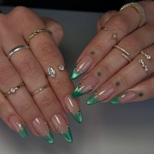 May include: A close-up of hands showcasing a set of long, almond-shaped nails with a French manicure design. The nails feature a nude base with a vibrant green tip, accented with gold detailing. Various gold rings adorn the fingers.