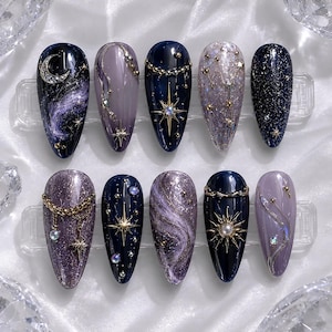 May include: A set of ten almond-shaped press-on nails featuring celestial designs in shades of deep navy, purple, and silver. The nails are decorated with metallic gold stars, moons, chains, pearls, and glitter, creating a cosmic night sky theme.