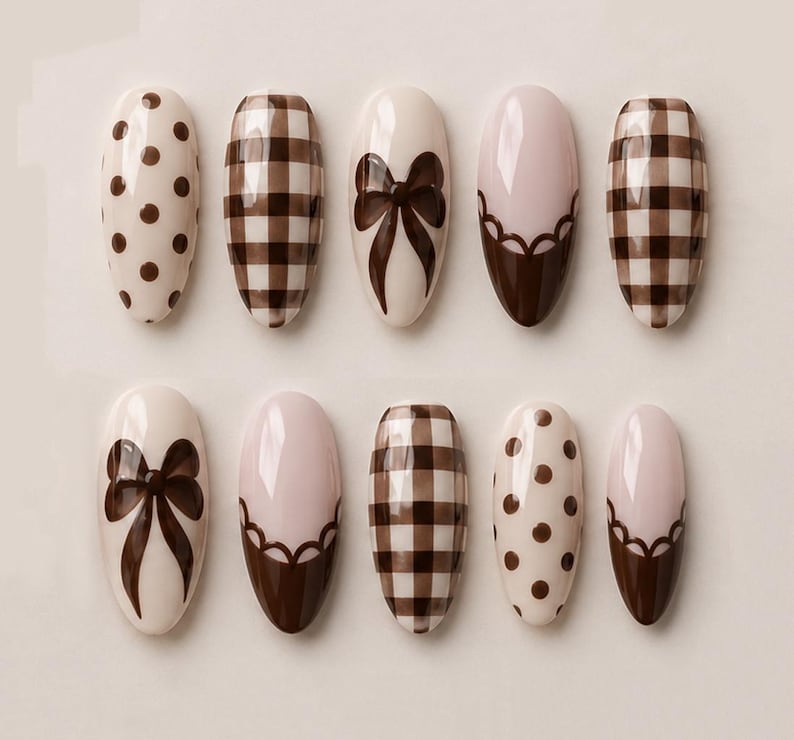 May include: A set of ten almond-shaped press-on nails featuring a cohesive brown and cream color palette. The designs include dark brown polka dots on a cream base, a brown and cream checkered pattern, solid brown bows, and scalloped brown tips.