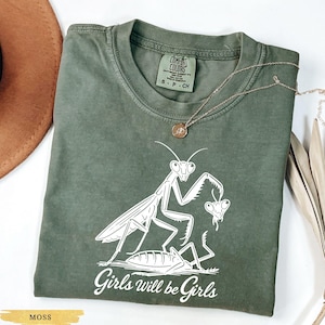May include: Moss green t-shirt with a white graphic of praying mantises and the text "Girls will be Girls." The shirt has a crew neck and short sleeves. A gold necklace is draped across the shirt.