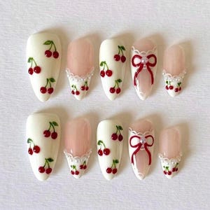 May include: A set of almond-shaped false nails. Some nails are painted white with red cherry designs and green leaves. Others are a pale pink with a white lace trim and small red cherries. One nail features a red bow.