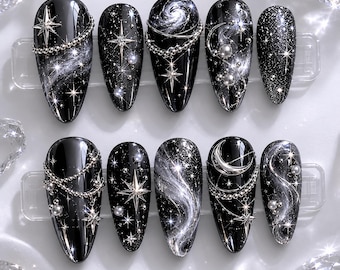 May include: A set of ten black press-on nails with a galaxy-inspired design. Each nail features silver glitter, stars, and swirling white accents. Some nails have silver chain and bead embellishments, creating a celestial theme.