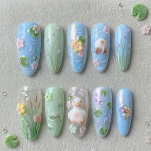 May include: A set of ten artificial nails with a spring theme. The nails are painted in pastel blue and green, with 3D floral and duck designs. Some nails feature water lily and cattail designs, with small rhinestones.