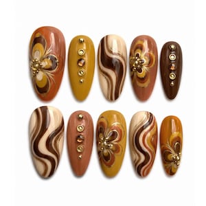 May include: A set of ten almond-shaped press-on nails featuring a color palette of brown, tan, and mustard yellow. The designs include swirling patterns, hand-painted floral motifs, and vertical rows of small gold-toned metallic studs and crystals.