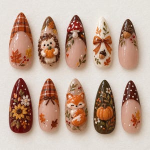 May include: A set of ten almond-shaped press-on nails featuring autumn-themed designs including a hedgehog, a fox, pumpkins, sunflowers, mushrooms, acorns, plaid patterns, and fall foliage in shades of orange, brown, red, and cream.