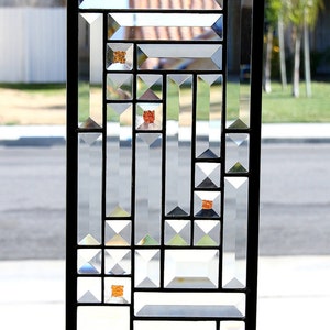May include: Rectangular stained glass window panel with a black metal frame. The panel features clear, geometric shapes and small orange accents. A chain is attached for hanging. The artist's signature is visible at the bottom.