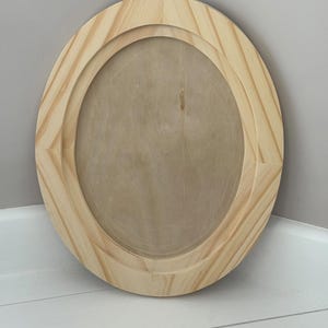 May include: Oval wooden picture frame with a natural wood finish. The frame has a layered design with a beveled edge. The interior oval is a lighter shade of wood. The frame is displayed in a corner against a gray wall and white floor.