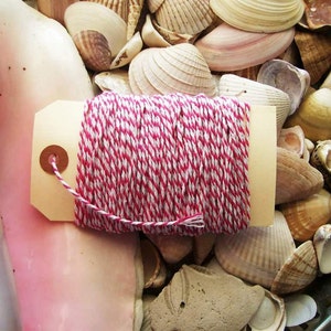 May include: A close-up of a spool of red and white baker's twine with a brown tag. The twine is sitting on a bed of seashells.