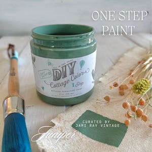 May include: A glass jar of green paint labeled "DIY Cottage Colors" with the text "All in One Paint" and "1 Step Artisan Furniture Paint 8 oz (236 ml)". The jar is sitting on a white wooden surface with a paintbrush and a piece of fabric with the text "Juniper" and "Curated by Jami Ray Vintage".