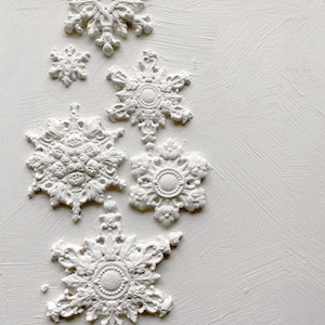May include: A collection of six white snowflake ornaments featuring intricate embossed floral and geometric patterns. Each piece displays a textured, matte finish with varying sizes and detailed radial symmetry, arranged in a vertical cluster.