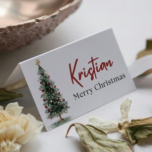 Peut inclure: Une carte de Noël blanche avec une illustration à l'aquarelle d'un sapin de Noël décoré. Le nom "Kristian" est écrit en script rouge, avec "Merry Christmas" en dessous. La carte est entourée de feuilles séchées et d'un bol décoratif.