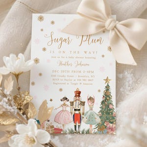 以下が含まれることがあります： 「Sugar Plum is on the way!」のテキストが入ったベビーシャワーの招待状。クリーム色のリボンと装飾的な花の配置が特徴です。招待状には、くるみ割り人形、バレリーナ、クリスマスツリーのイラストが含まれています。