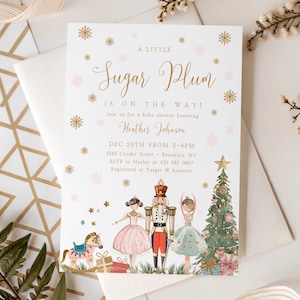 Op de afbeelding: Een uitnodiging voor een babyshower met de tekst "A Little Sugar Plum Is On The Way!" in gouden script. De uitnodiging bevat illustraties van een notenkraker, ballerina's, een kerstboom en een schommelpaard. Roze en gouden accenten.