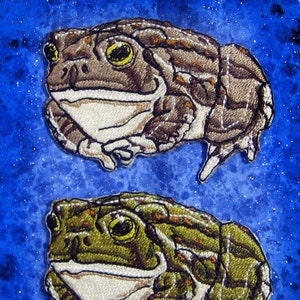 May include: Two embroidered patches of toads on a blue background with white speckles. The toad on top is brown with a white belly and the toad on the bottom is green with a white belly.