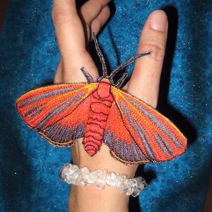 Peut inclure: Un mannequin de main portant un bracelet avec un motif de papillon de nuit brodé rouge, noir et orange.