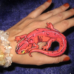 May include: A red embroidered patch of a salamander with black accents. The patch is on a person's hand.