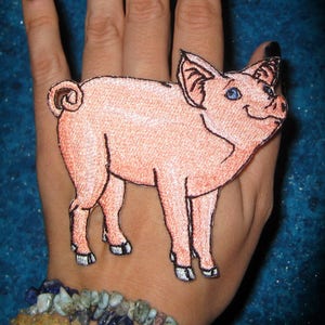 Op de afbeelding: Een roze varkens geborduurde patch met blauwe ogen. De patch zit op de hand van een persoon met een armband om de pols.
