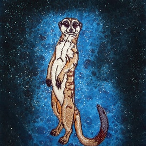 May include: Embroidered patch featuring a meerkat standing upright. The meerkat has tan and brown fur with a white chest and a dark tail. The background is a blue nebula with white stars, creating a space-themed design.