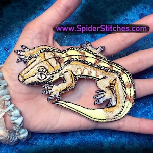 May include: An embroidered gecko patch in yellow and brown with a white background. The patch is on a hand wearing a bracelet made of colourful stones. The text "www.SpiderStitches.com" is visible in the background.