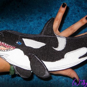 以下が含まれることがあります： 黒いフェルトと白いフェルトでできた、口を開けて白い歯と青い目をしたシャチのアップリケ。アップリケは人の手につけられています。