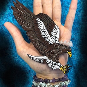 May include: Brown and white embroidered eagle patch with yellow talons. The patch is on a hand with a blue background.