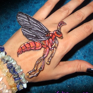 May include: A red and black embroidered iron-on patch of a wasp with wings spread. The patch is on a person's hand with black nail polish.