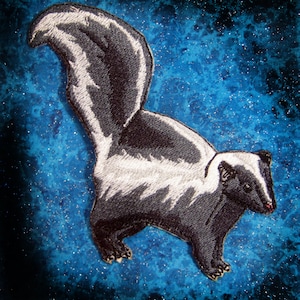 Könnte beinhalten: Ein gestickter Aufnäher mit einer schwarz-weißen Stinktier-Darstellung, die einen weißen Streifen auf dem Rücken hat. Der Aufnäher befindet sich auf einem blauen Hintergrund mit weißen Punkten.