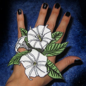 May include: A white embroidered patch of two datura flowers with green leaves. The patch is on a hand with black nail polish.