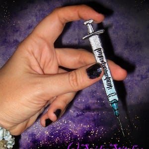 May include: A hand holding a silver glitter syringe with black ink detailing. The syringe is held against a purple background with gold glitter.