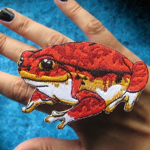 May include: Red and yellow embroidered frog patch with white accents. The patch is on a hand with black nail polish.