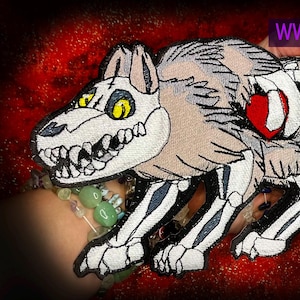 Puede incluir: Parche bordado de un esqueleto de lobo con ojos amarillos y un corazón rojo. El parche es de color beige, negro y blanco, con el texto "WWW.SpiderStitches.com" en morado.