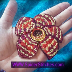 May include: A red, yellow, and purple embroidered flower patch with a striped pattern. The patch is on a hand with a bracelet.