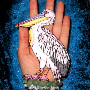 May include: A pink and white embroidered pelican patch with black accents. The patch is on a hand wearing a multi-colored beaded bracelet.
