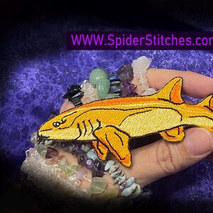 Puede incluir: Parche bordado con un tiburón amarillo y naranja estilizado. El parche tiene costuras detalladas y se sostiene sobre un fondo texturizado. La dirección del sitio web "WWW.SpiderStitches.com" se muestra en una pancarta púrpura.