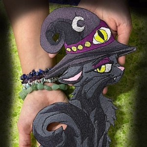 May include: Embroidered patch featuring a black cat wearing a witch's hat. The hat is purple with a yellow and purple band. The cat has yellow eyes and pink accents. The patch is held in hands. The website address www.SpiderStitches.com is at the bottom.