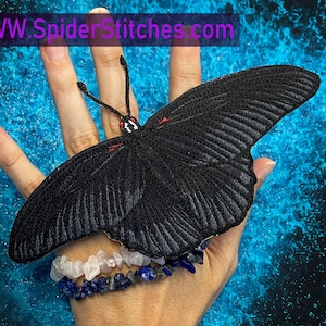 Könnte beinhalten: Ein schwarzer gestickter Schmetterlings-Patch mit roten Akzenten, gehalten in einer Hand, die ein Armband mit weißen und blauen Steinen trägt. Der Hintergrund ist eine blau-schwarze gesprenkelte Oberfläche. Der Text "WWW.SpiderStitches.com" ist oben im Bild sichtbar.