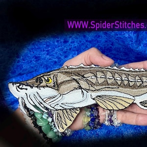May include: Embroidered patch of a sturgeon fish in shades of brown, tan, and white, with yellow eye details. The patch is held in a hand against a blue background. Text on the image reads "WWW.SpiderStitches.com".