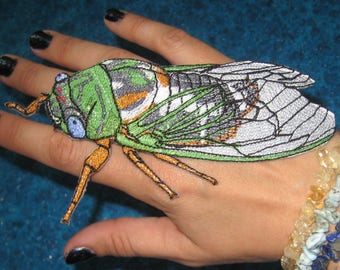 May include: A green and white embroidered cicada insect patch with brown legs and a white background. The patch is on a person's hand wearing a multi-colored beaded bracelet.