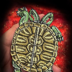May include: An embroidered patch depicting a two-headed turtle, with green and brown colouring and red markings on its head. The turtle is facing upwards with its shell visible. The patch is set against a red background with white text overlaying it, reading "WWW.SpiderStitches.com".