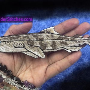 May include: A brown and white embroidered shark patch with a blue eye. The patch is about 6 inches long and is held in a hand. The text "WWW.SpiderStitches.com" is visible in the top left corner of the image.