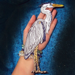 May include: A gray and white embroidered heron patch with yellow legs and a long, thin beak. The patch is on a person's hand, with a blue and green background.