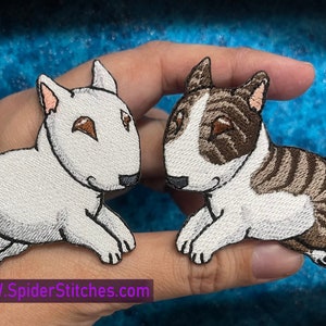 May include: Two embroidered patches of white bull terrier puppies. One patch is white with brown eyes and the other is white with brown stripes and brown eyes. The patches are on a hand.