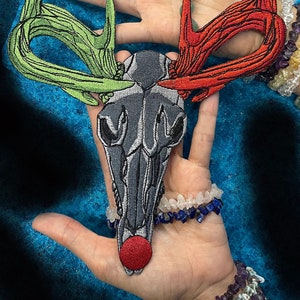 May include: A gray embroidered deer skull with red and green antlers. The skull has a red nose and the antlers are stitched with a detailed design. The skull is held in a hand with a bracelet on the wrist.
