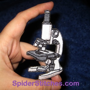 May include: White and black embroidered iron-on patch of a microscope. The patch is about 2 inches tall and 1.5 inches wide. The patch is on a blue background.