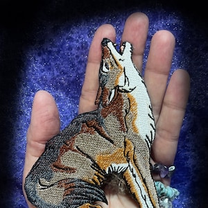 May include: A brown and white embroidered patch of a wolf howling at the moon. The patch is on a hand with a bracelet of colorful stones. The text "WWW.SpiderStitches.com" is at the bottom of the image.