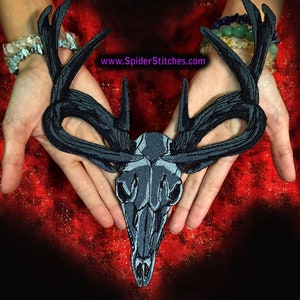 Puede incluir: Un parche bordado en negro y gris que representa una calavera de ciervo con cuernos. El parche está en las manos de una persona. El texto "www.SpiderStitches.com" es visible en la imagen.