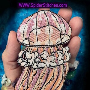 May include: Embroidered jellyfish patch in shades of pink, orange, and white, outlined in black. The jellyfish has a domed top with vertical stripes and flowing tentacles. The text "WWW.SpiderStitches.com" is visible at the top.